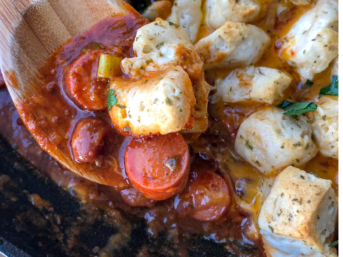 A wooden spoonful of chili, hot dogs, green onions, and garlic butter biscuits in a cast iron skillet.