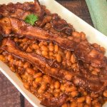 White oval serving dish of baked beans and strips of bacon.