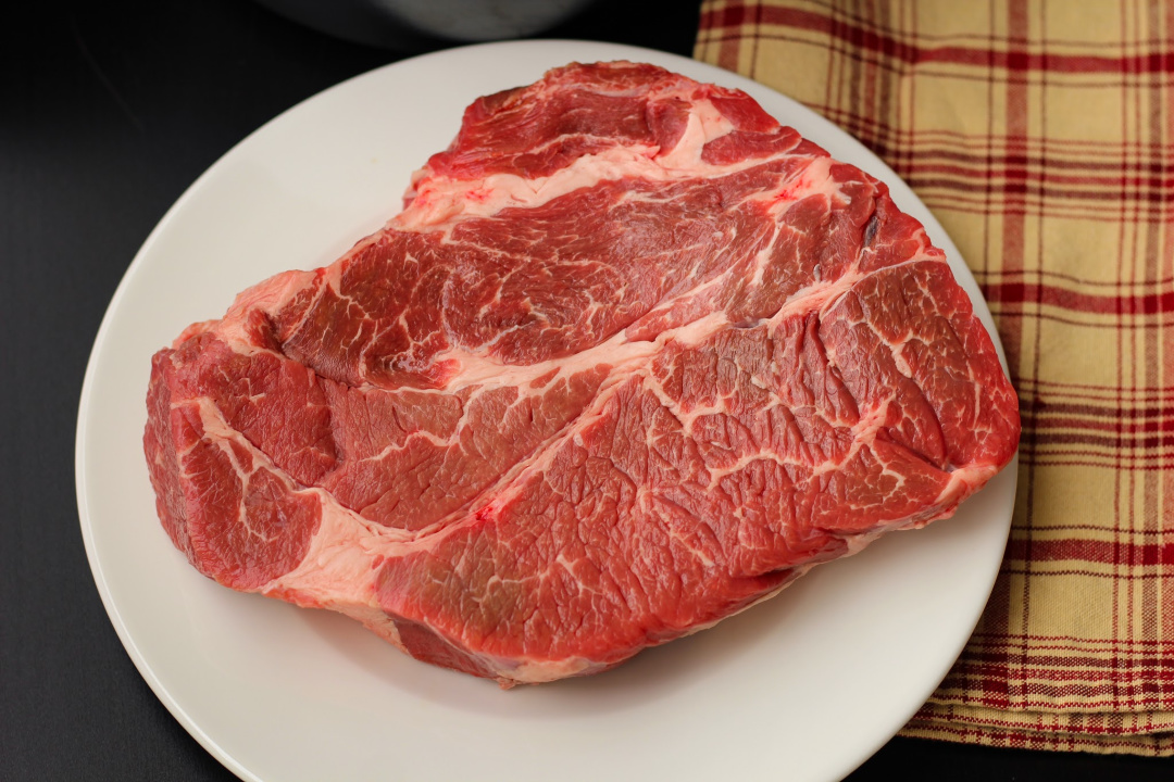 A 2.78 pound of chuck roast with perfect marbling on a white plate before being seasoned and seared.