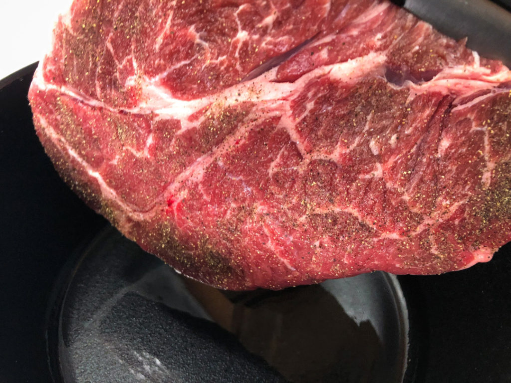 The seasoned chuck roast is being placed in a hot dutch oven with cooking oil to sear it.