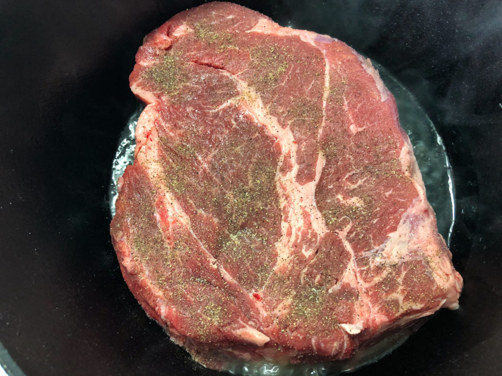 Seasoned chuck roast being seared in a dutch oven over medium high heat. 