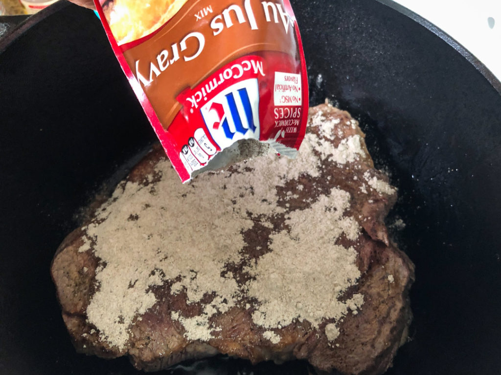 The packet of Au Jus Gravy mix is being sprinkled over the seared chuck roast in the Dutch oven.