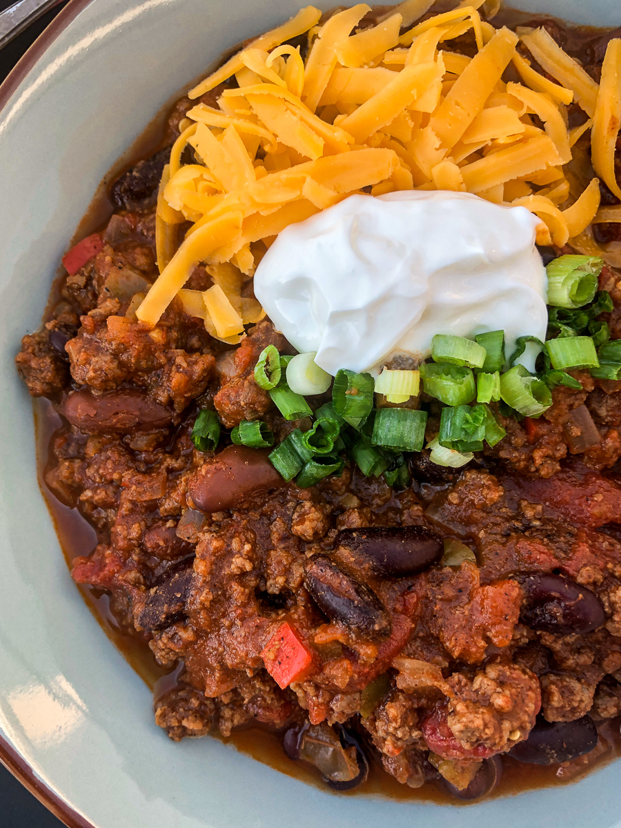 Close up and side view of Awesome Chili in a blue-gray bowl garnished with sour cream, green onions, and shredded cheddar cheese. 