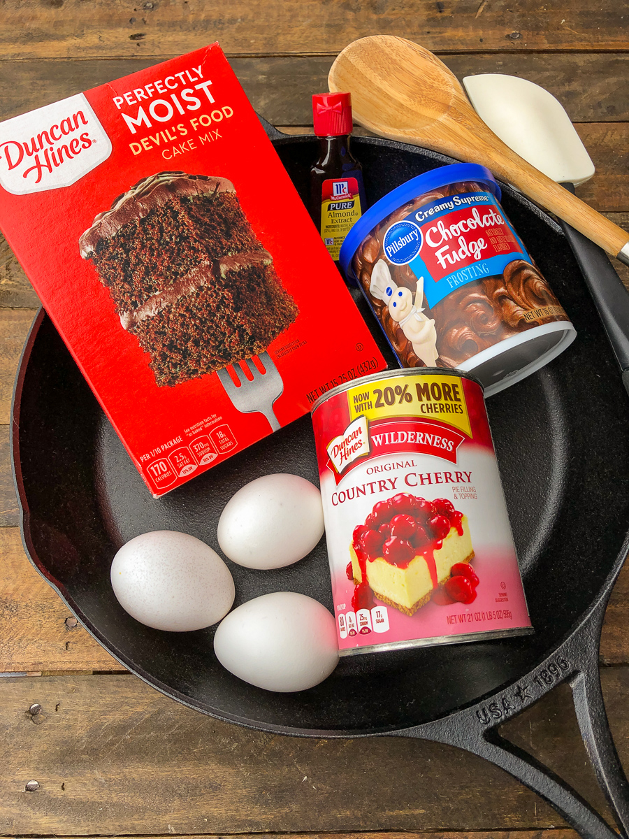 Chocolate Cherry Cake ingredients, including the cake mix, almond extract, eggs, cherry pie filling all in the cast iron skillet with a wooden spoon and rubber spatula. 