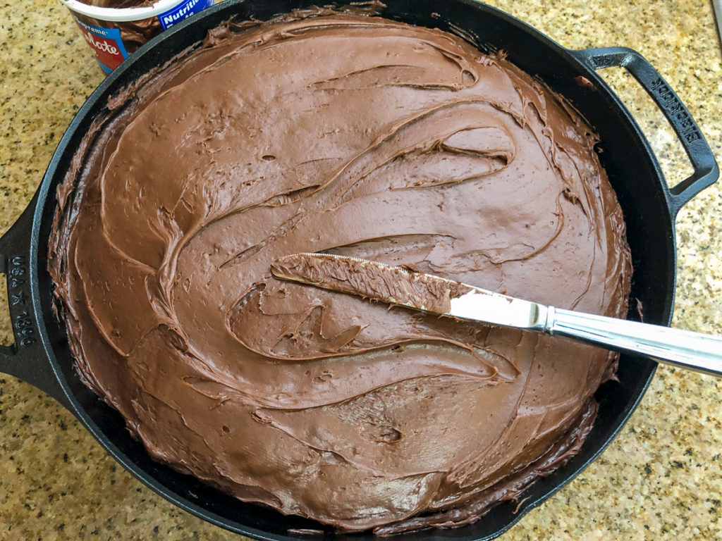 The freshly baked chocolate cherry cake has been frosted while still in the skillet with a butter knife in view. 