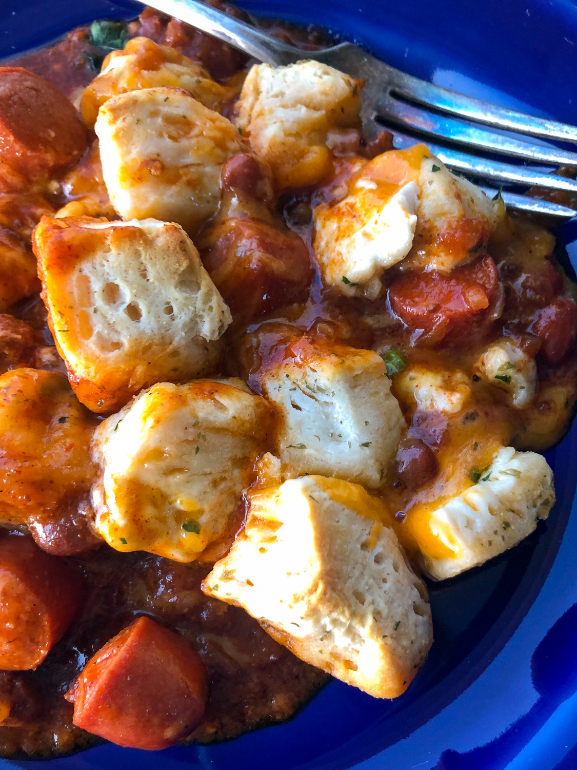 Pillowy biscuits covered in garlic butter over a mixture of chili, green onions, hot dogs, and cheddar cheese, on a blue plate. 