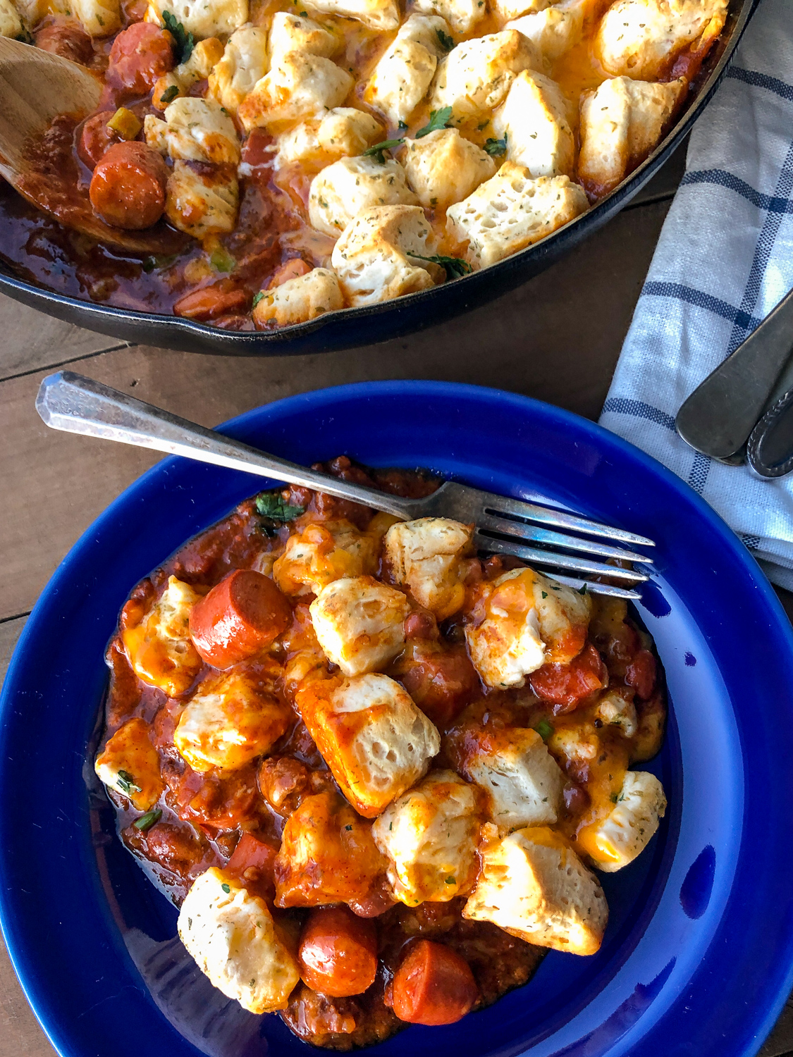 Cast iron chili cheese dog skillet bake in the background and a blue plate full of the cooked and served meal. 