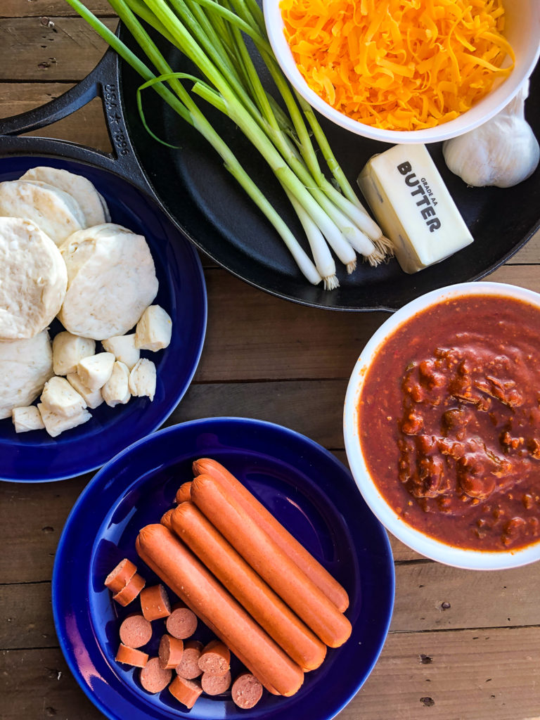 Ingredients to make this recipe: Hot dogs, chili, canned biscuits, green onions, butter, garlic, and cheddar cheese along with a cast iron skillet. 