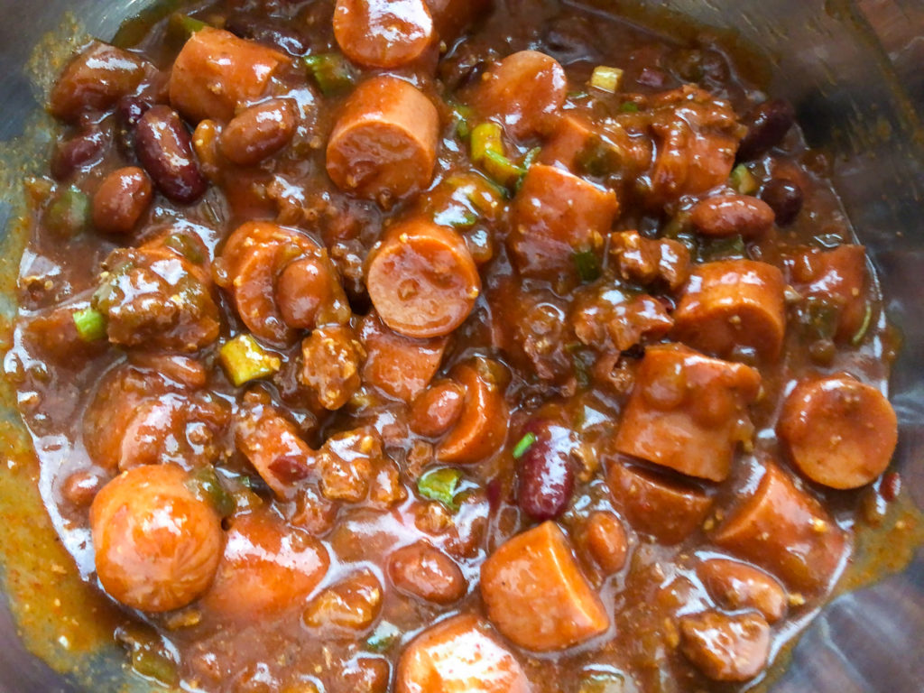 Sliced hot dogs and green onions have been added to the chili in a stainless steel bowl. 