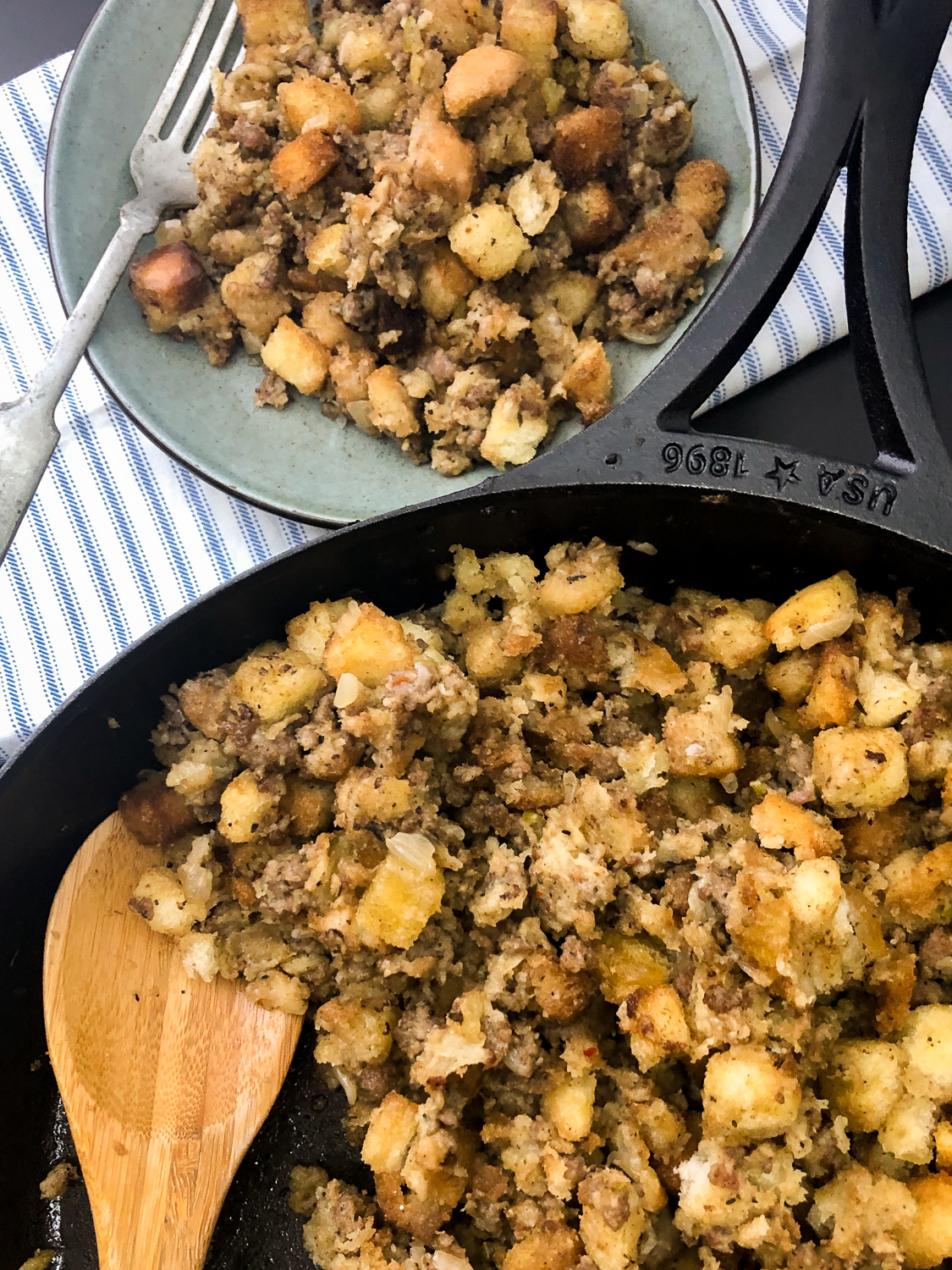 Cooked and served sausage stuffing portioned onto a blue plate with a fork and the rest remaining dressing in the skillet with a wooden spoon. 