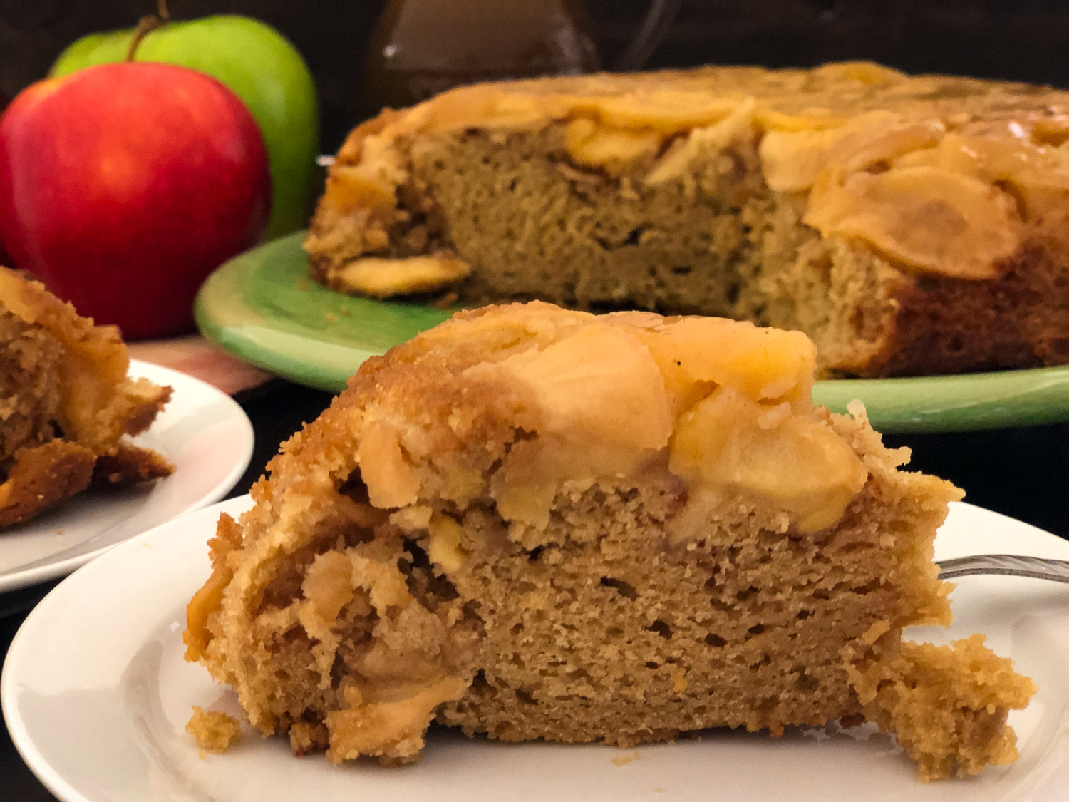 Slice of apple cake on a white plate with another slice and the remaining platter in the background.