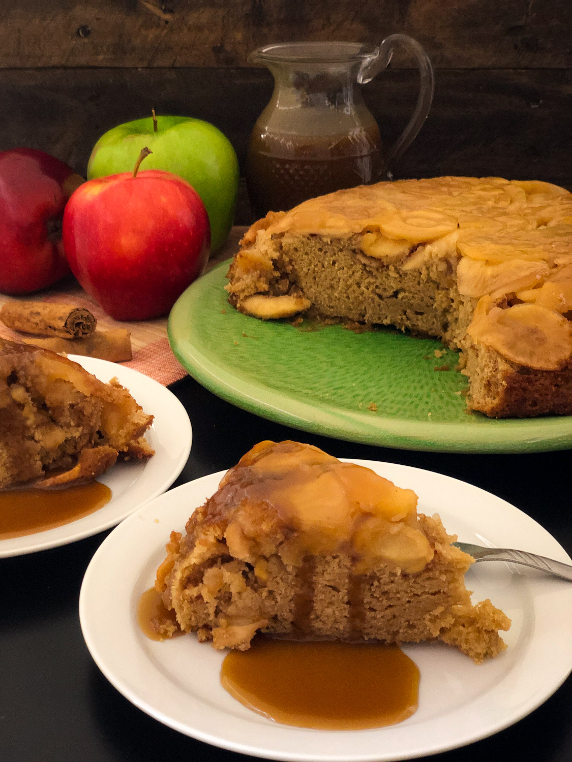 Slices of Dutch oven apple cake on white plates with caramel  sauce poured over; remaining cake on green  platter with pitcher of caramel sauce in the background.