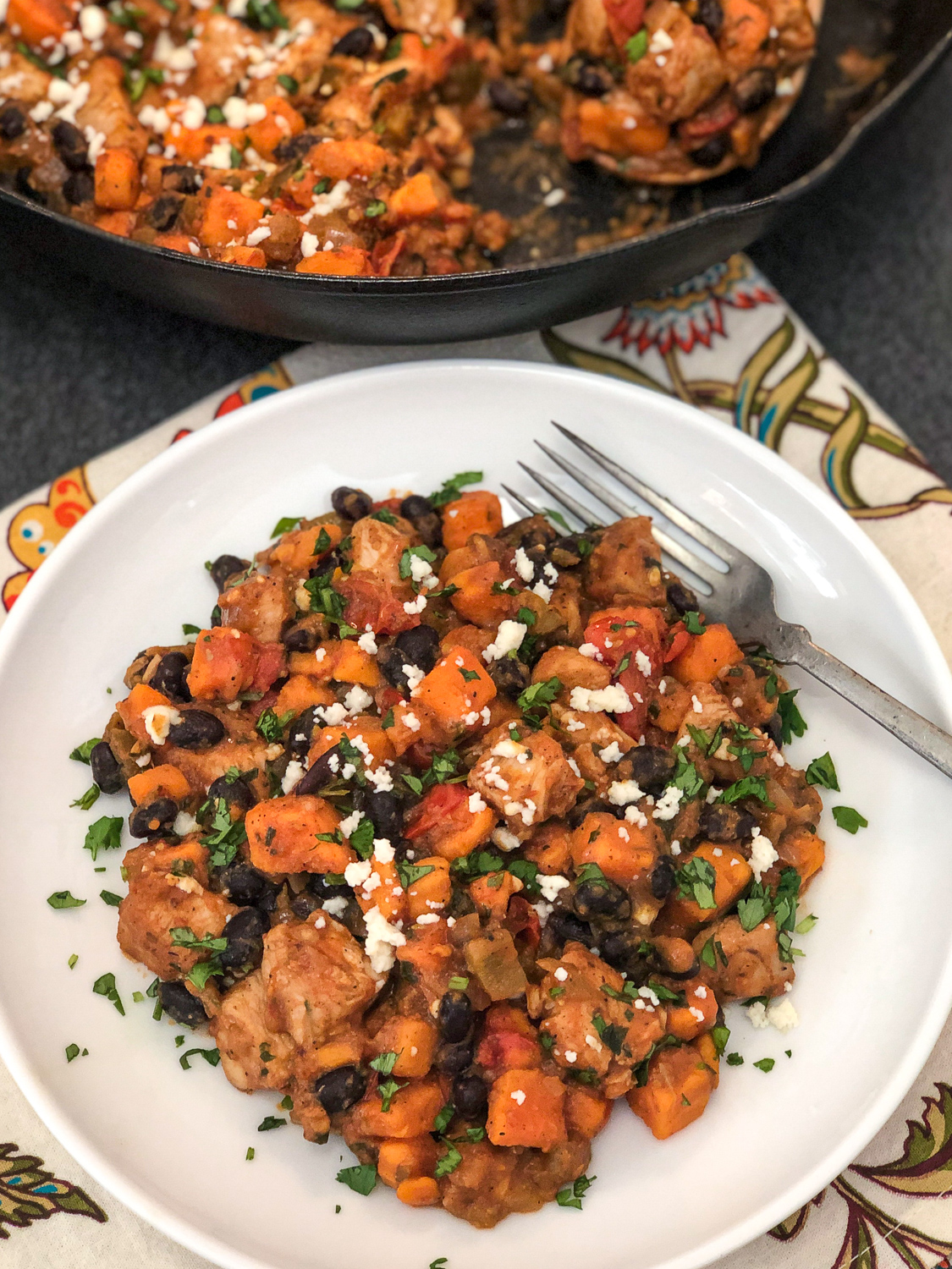 A serving of the skillet dinner on a white plate with a fork and a bright floral napkin; cast iron skillet in the background. 