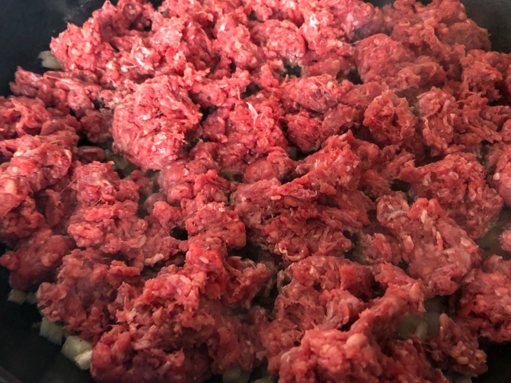 Ground beef crumbled into a cast iron skillet over sautéing onions. 