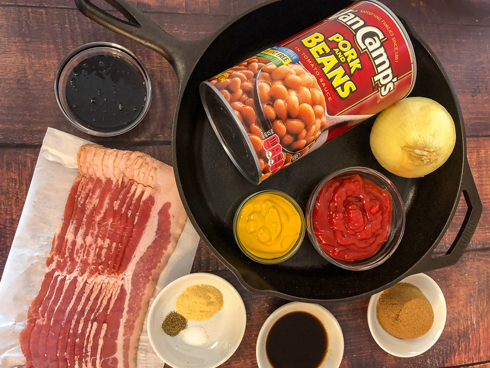 Ingredients to make baked beans with bacon: dark brown sugar, Worcestershire sauce, seasonings, bacon, molasses, pork and beans, onion, ketchup, and mustard with a cast iron skillet