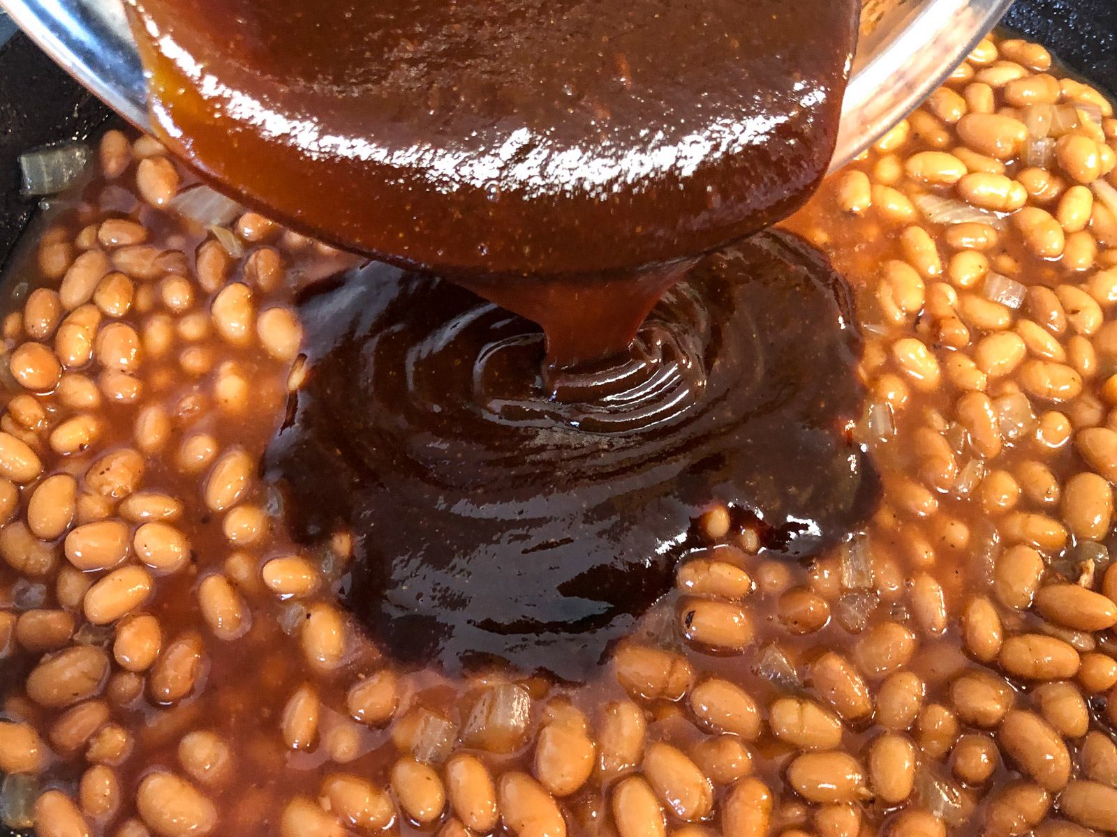 Homemade sauce being poured over pork and beans in a cast iron skillet