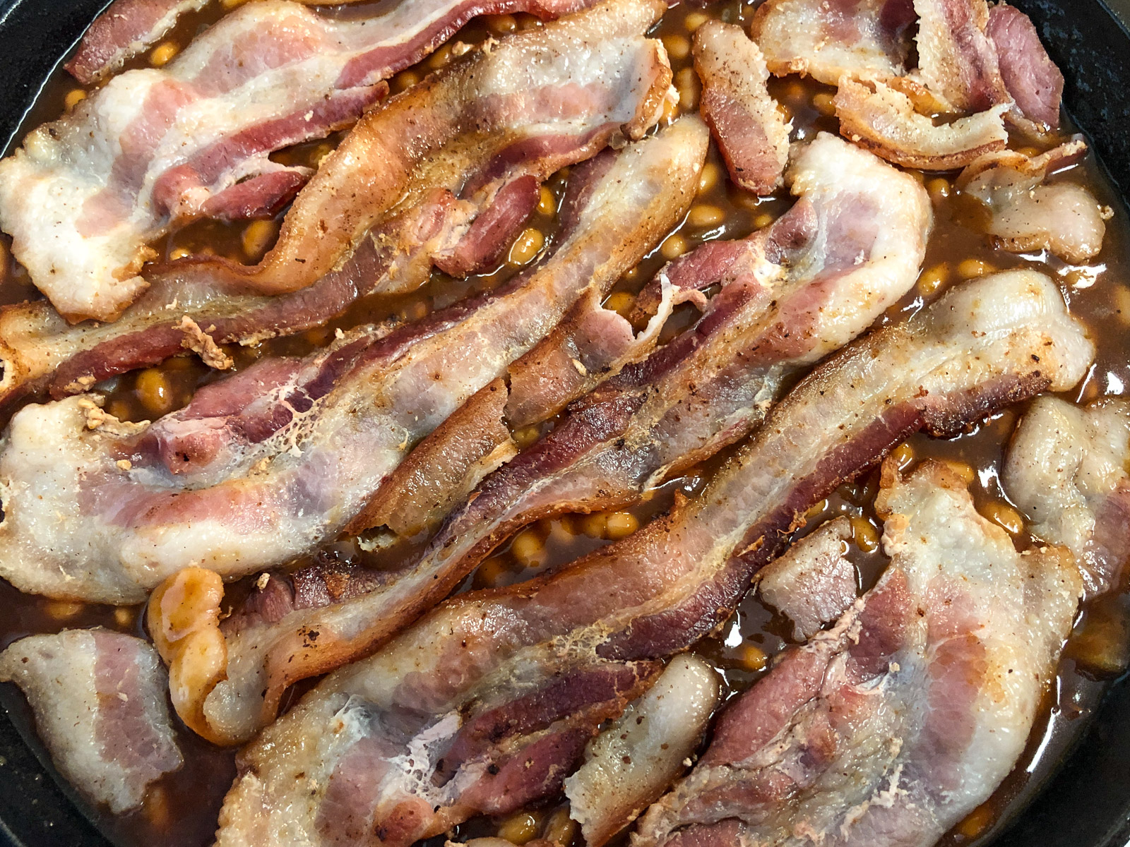 Bacon strips laid across the top of baked beans in a cast iron skillet ready for the oven