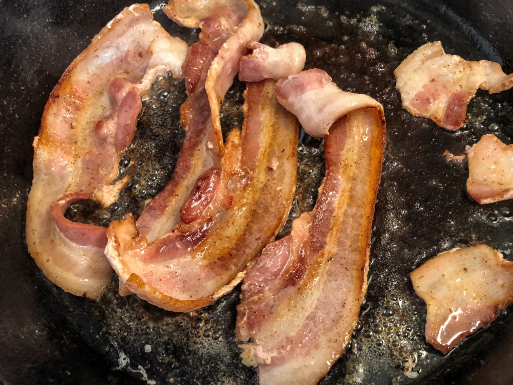 Strips and pieces of bacon cooking in a cast iron skillet.