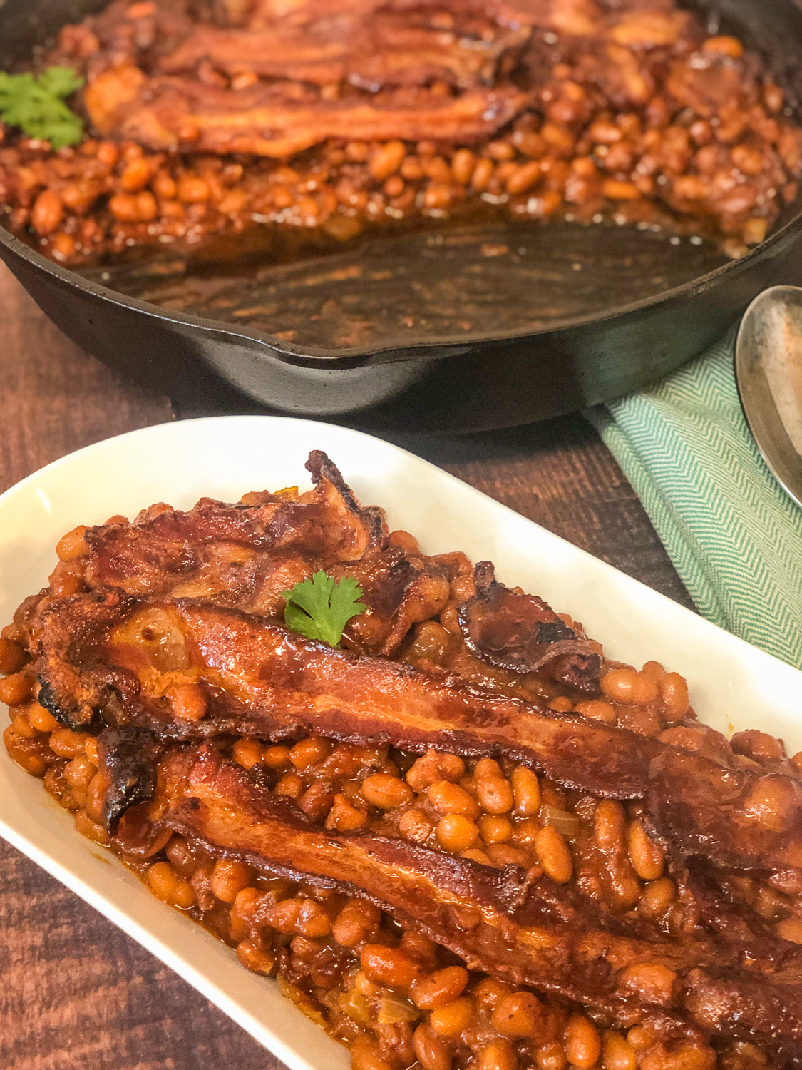 An oval white platter of baked beans topped with bacon; the cast iron skillet in the background with the remaining beans.