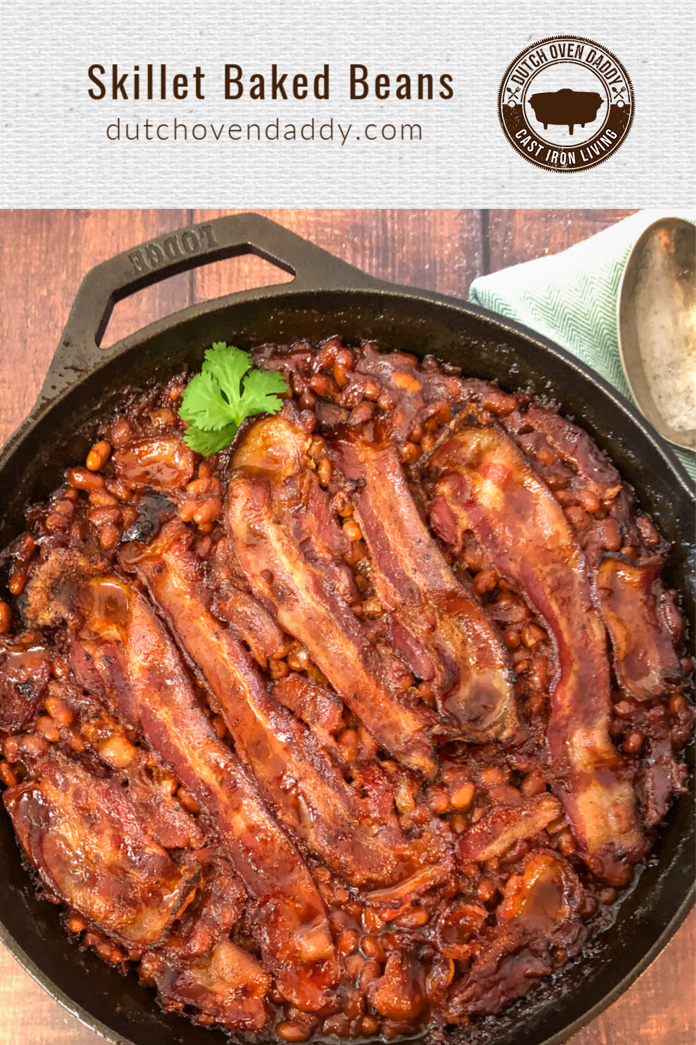 Cast iron skillet of baked beans topped with bacon strips and a green garnish, a green napkin and serving spoon off to the side.