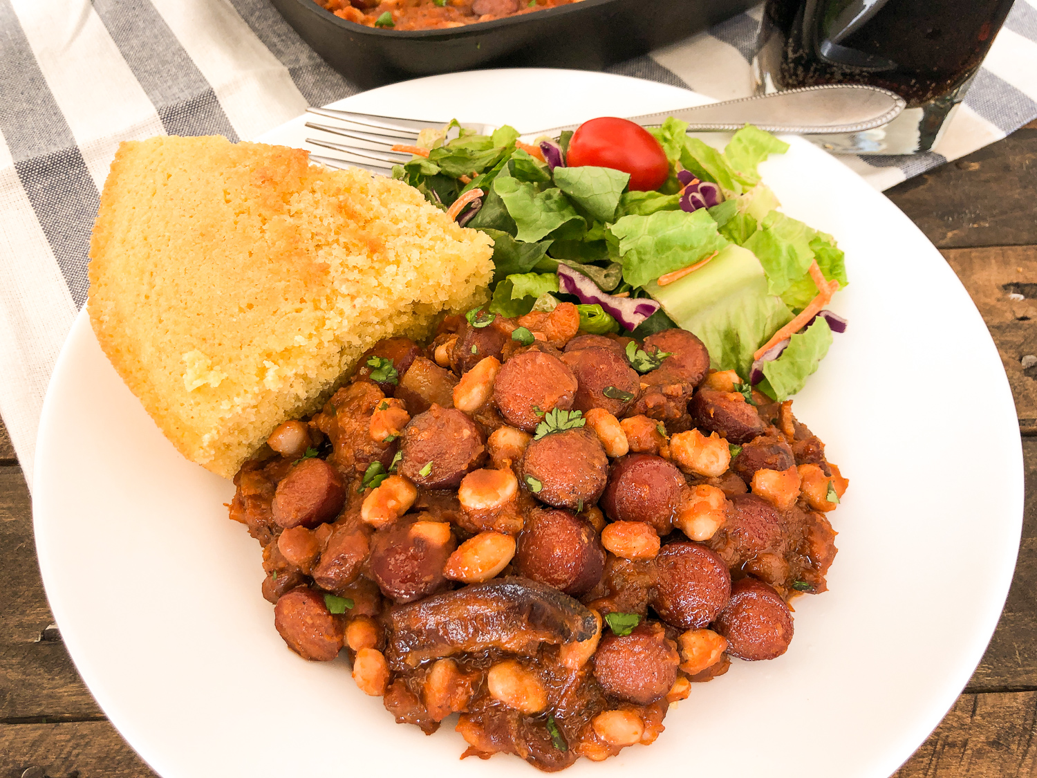 Cornbread, salad, and beans n franks on a white plate, a glass of soda and the skillet nearly out of frame.