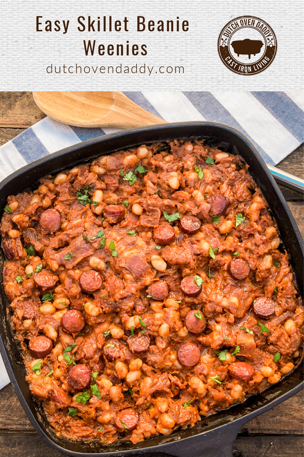 Square cast iron skillet filled with sliced hot dogs mixed into baked beans; wooden spoon resting on a blue and white linen. 