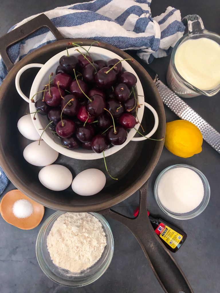 Ingredients needed to make the recipe in and around a cast iron skillet: salt, flour, almond extract, sugar, lemon zest, cream, eggs, and fresh cherries