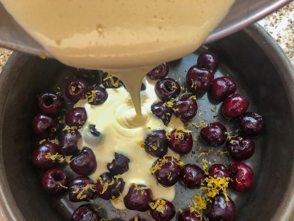 The clafoutis batter is being poured over cherries and lemon zest in a cast iron skillet. 