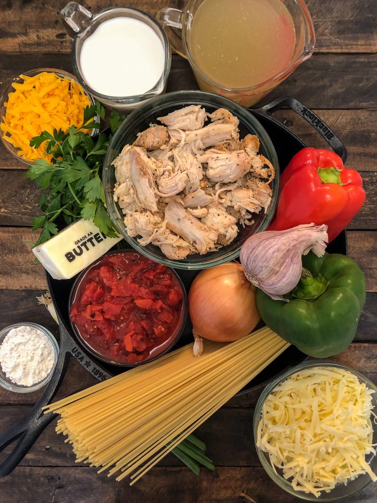 Ingredients needed to make chicken spaghetti in and around a cast iron skillet: cheddar cheese, cream, broth, chicken, parsley, butter, chicken, bell peppers, garlic, onions, diced canned tomatoes, flour, green onions, and white cheddar cheese. 