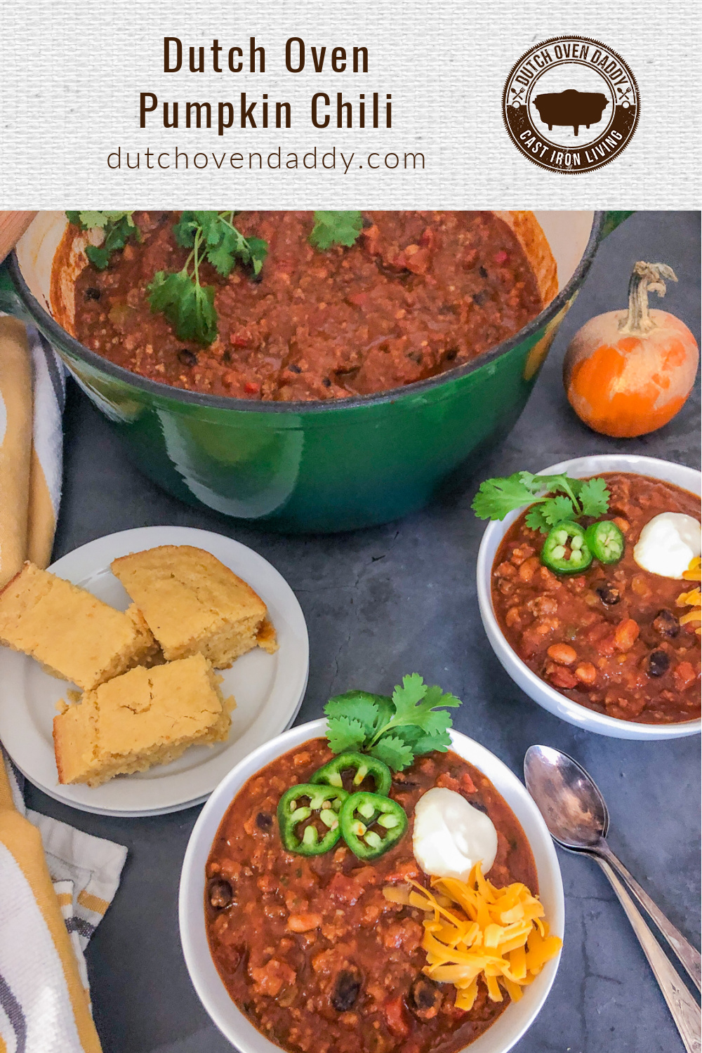 Branded image of Dutch Oven Pumpkin Chili showing it in the dutch oven, and served two white bowls, garnished with cheese, sour cream, jalapeños, and cilantro and a plate of cornbread off to the side. 