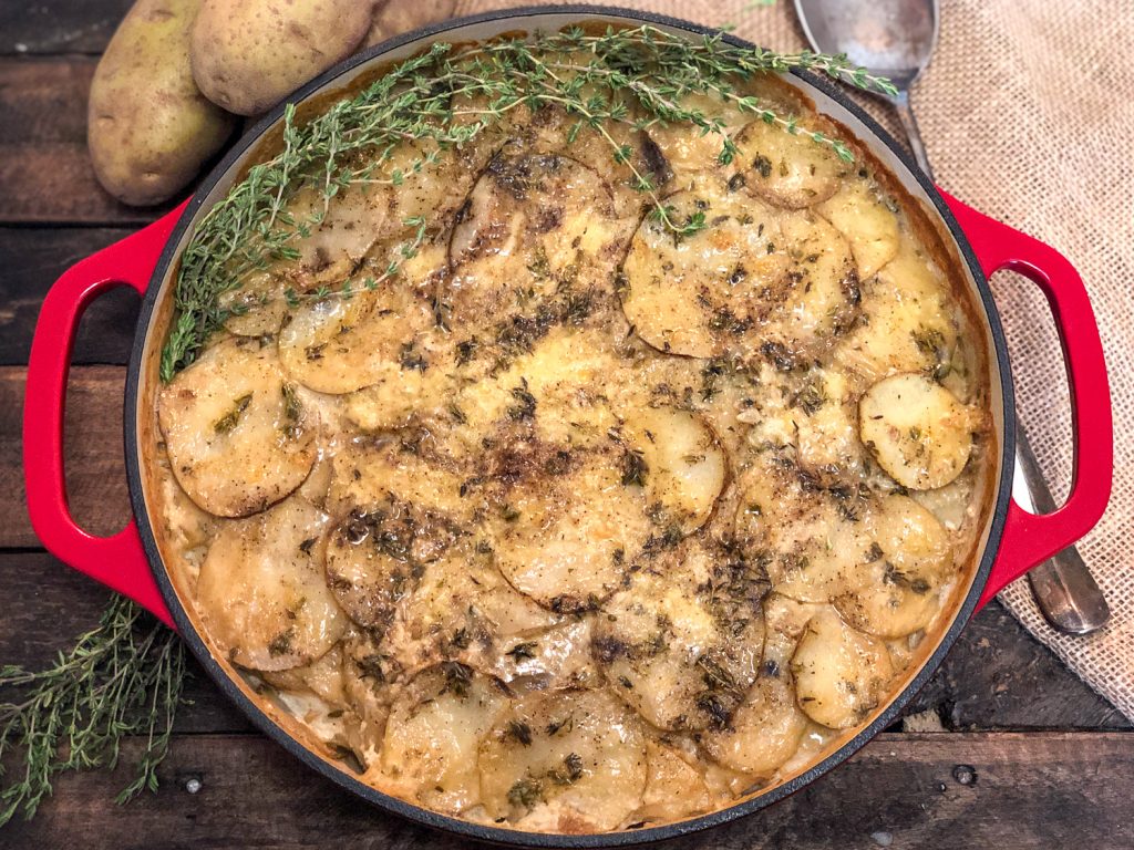 Freshly baked and rested Skillet Scalloped Potato Gratin garnished with fresh thyme sprigs, and potatoes and a serving spoon off to the sides.