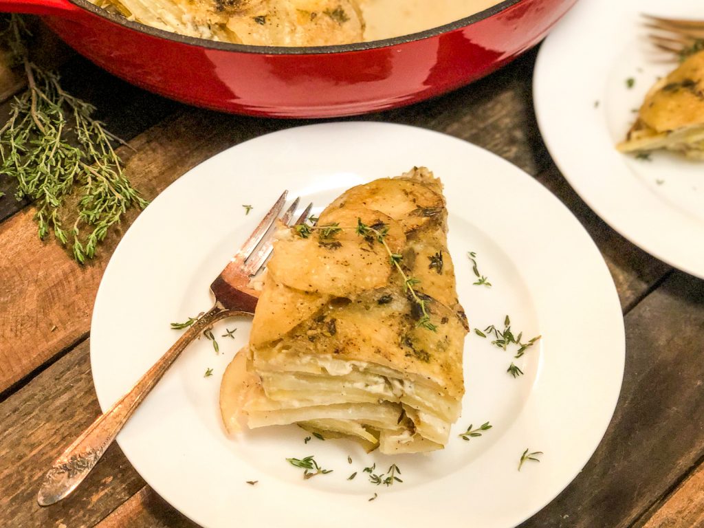 A serving of Skillet Scalloped Potato Gratin on a white plate with a fork. The red skillet in the background and fresh thyme garnishing the potatoes and in the background.