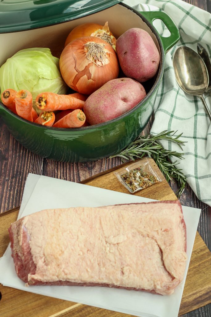 A green enameled Dutch oven with a cabbage, onions, and potatoes inside; corned beef on a cutting board with the spice packet; fresh rosemary, serving utensil and green/white dish cloth off to the side.