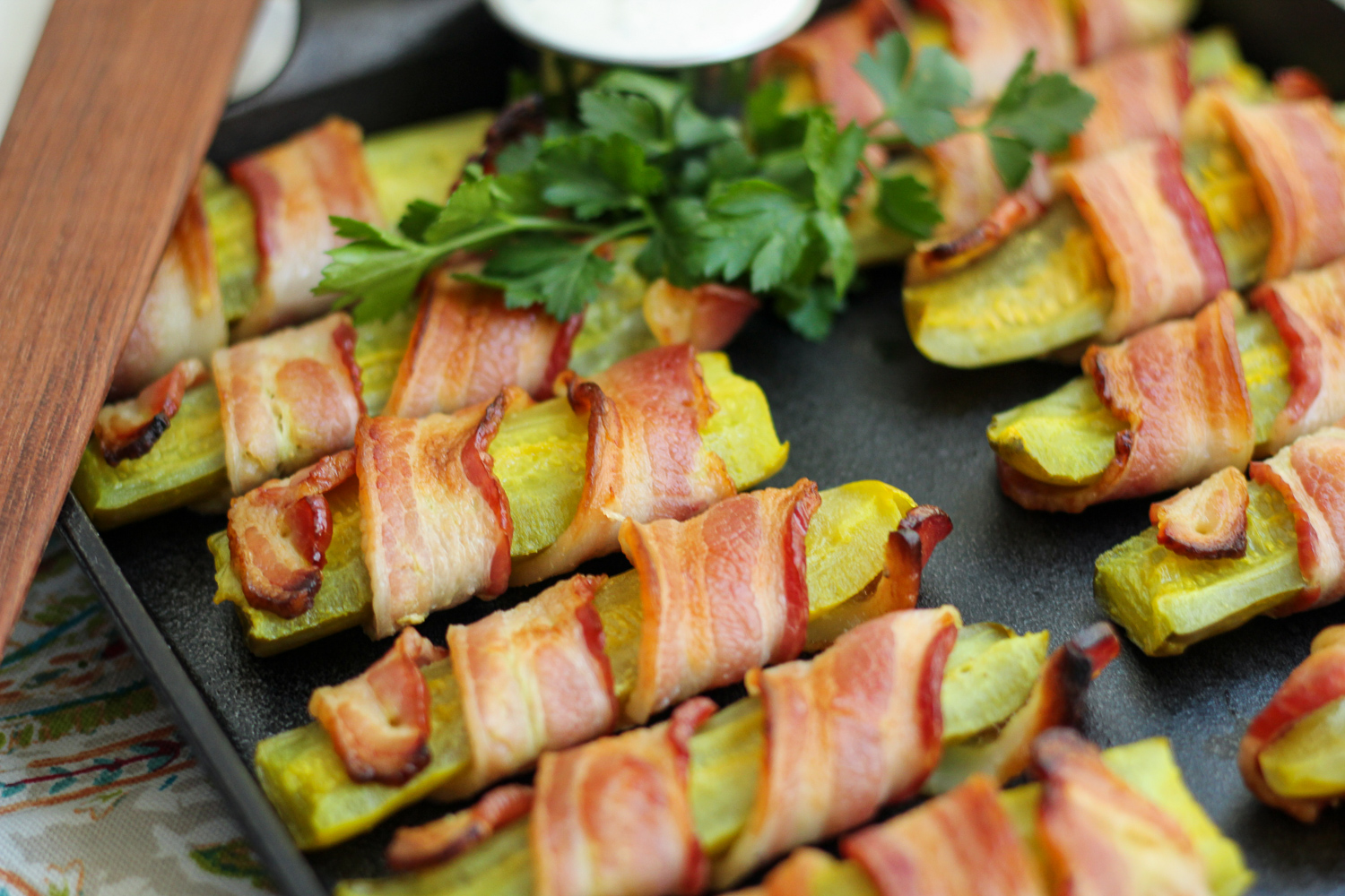 Bacon wrapped pickles on the cast iron baking pan garnished with fresh parsley and ranch dip.