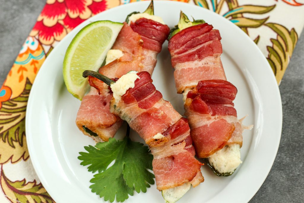 A white plate of 3 bacon wrapped jalapeño poppers with a lime wedge. 