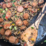 Skillet of Sausage, Peppers, and Rice with a portion missing and a handmade wooden spoon.