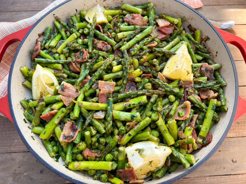The completed dish of peas, asparagus, bacon, leeks, garden herbs, and lemon in a red enameled Dutch oven.