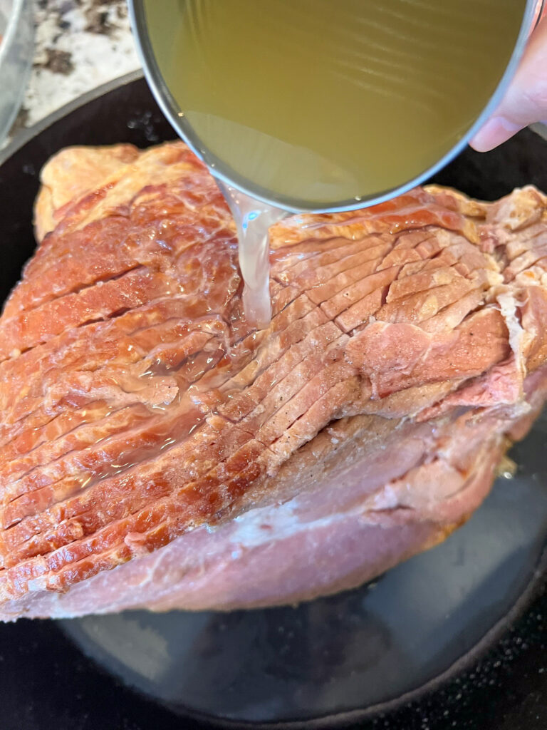 Broth being poured over the scored ham in the cast iron skillet.