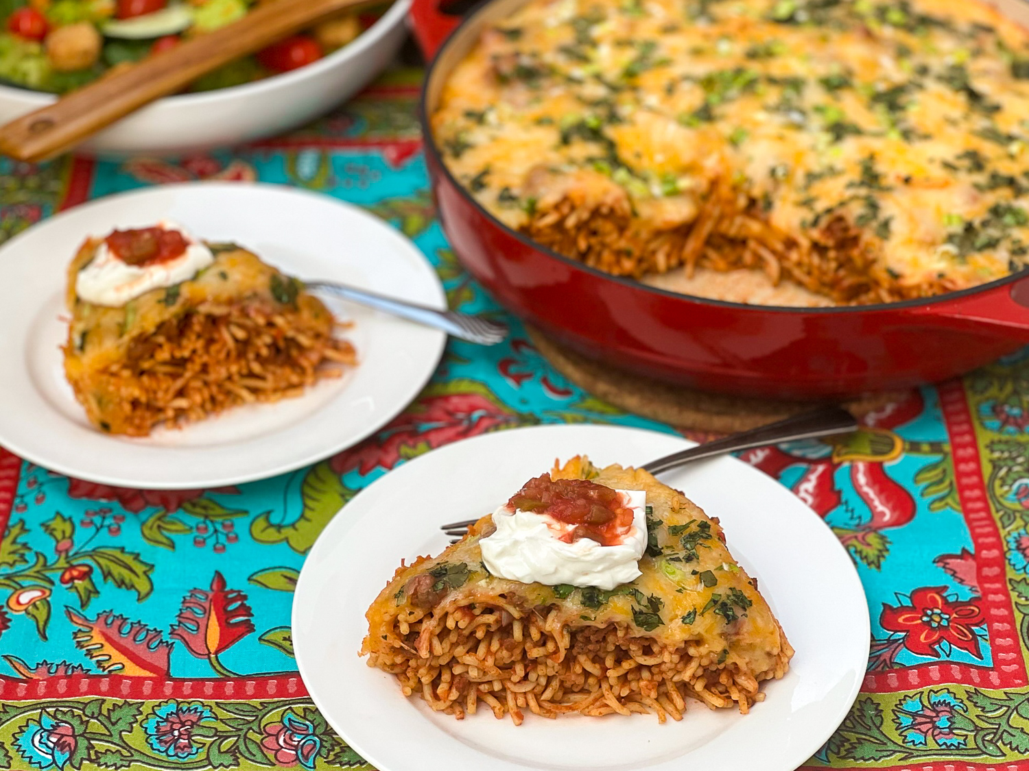 Two slices of Baked Mexican spaghetti on white plates with a salad and the remaining pasta in the skillet in the background.