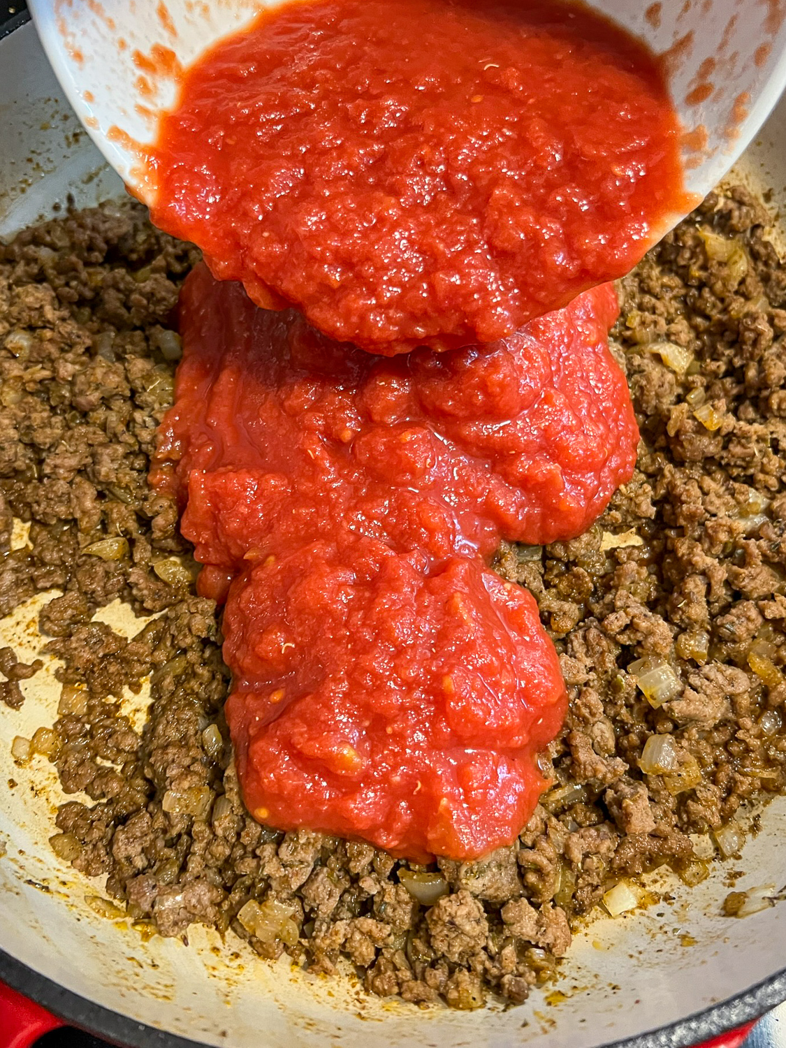 Crushed tomatoes being added to the seasoned cooked meat mixture.