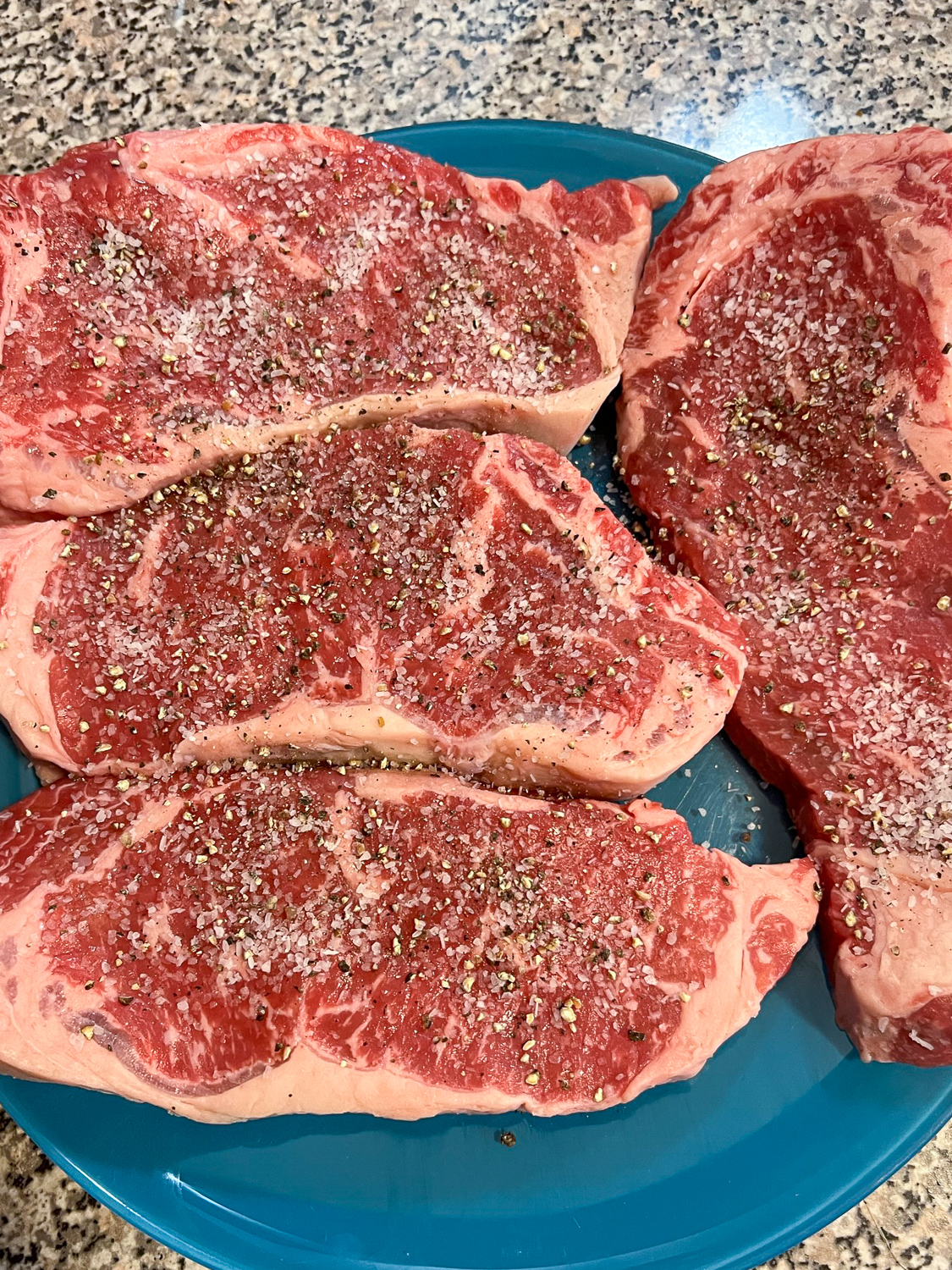 4 New York strip steaks on a plate coming to room temperature seasoned with salt and pepper.