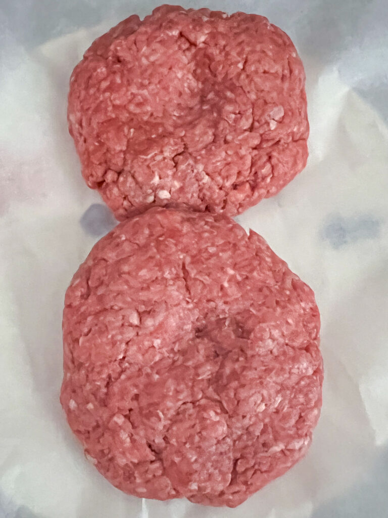 Two half pound patties seasoned and formed sitting on wax paper on a plate.