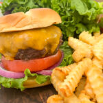 Cheeseburger in a bun with lettuce, tomato, and red onion sitting next to french fries.