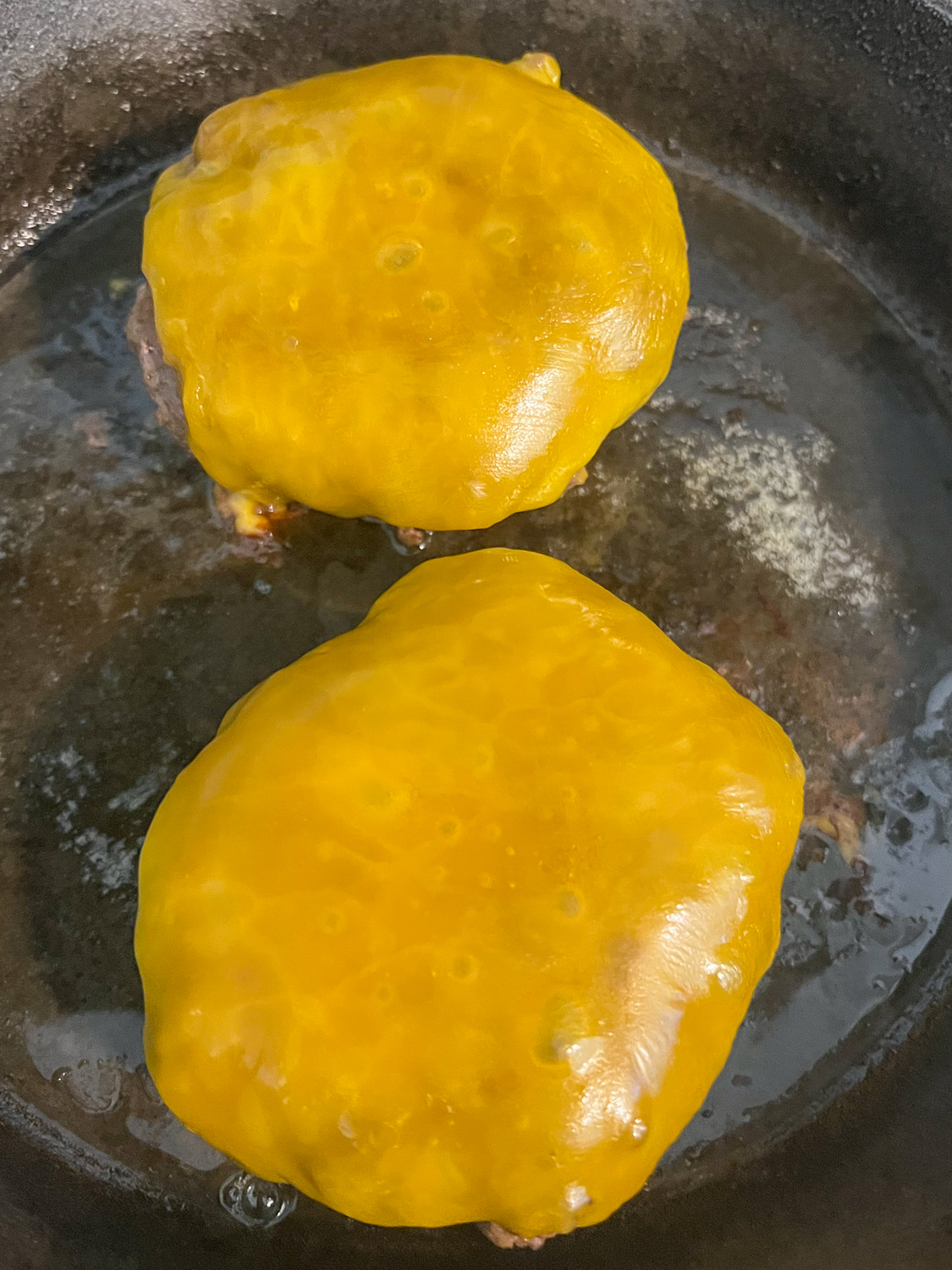 Two hamburger patties with melted cheddar cheese in a cast iron skillet.