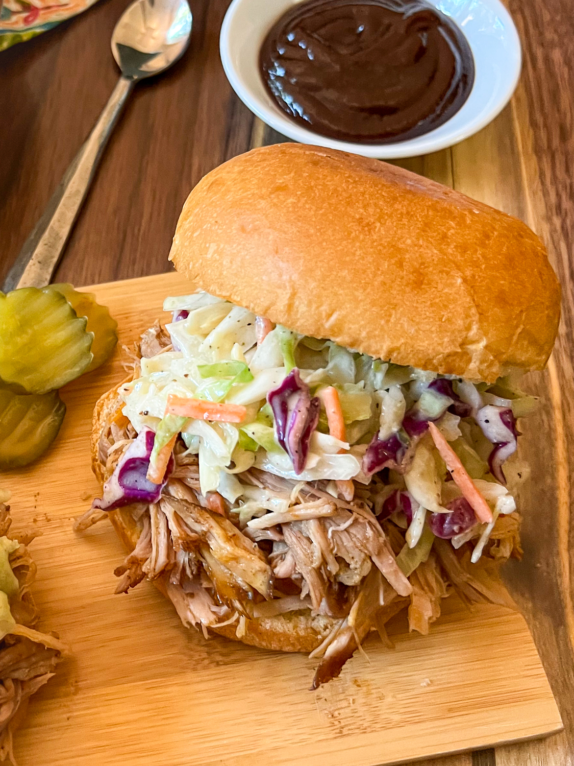 Close up view of the pulled pork in a bun topped with coleslaw; pickles off to the side and bbq sauce in the background. 