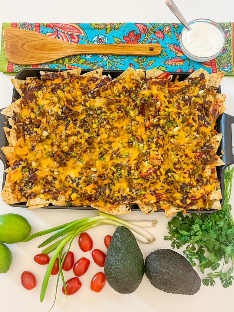 Fresh from the oven platter of pulled pork nachos with garnishes of lime, tomatoes, green onion, sour cream, avocados, and cilantro surrounding it.