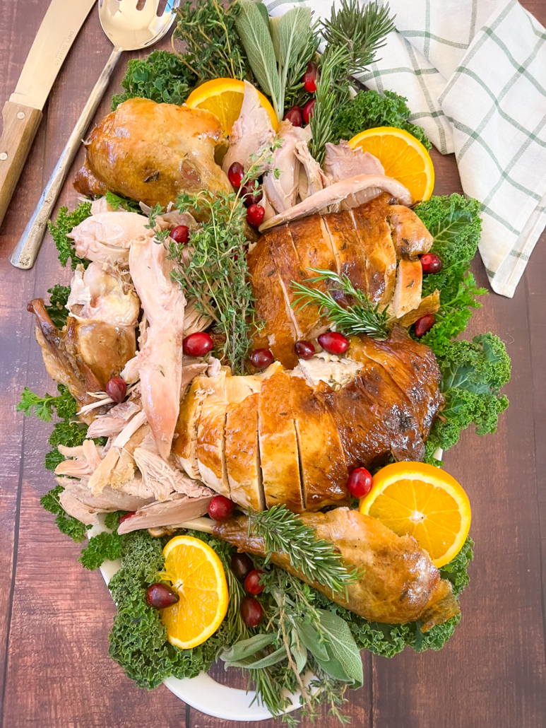 The whole turkey carved and plated on a white platter garnished with fresh herbs, cranberries, and orange slices.