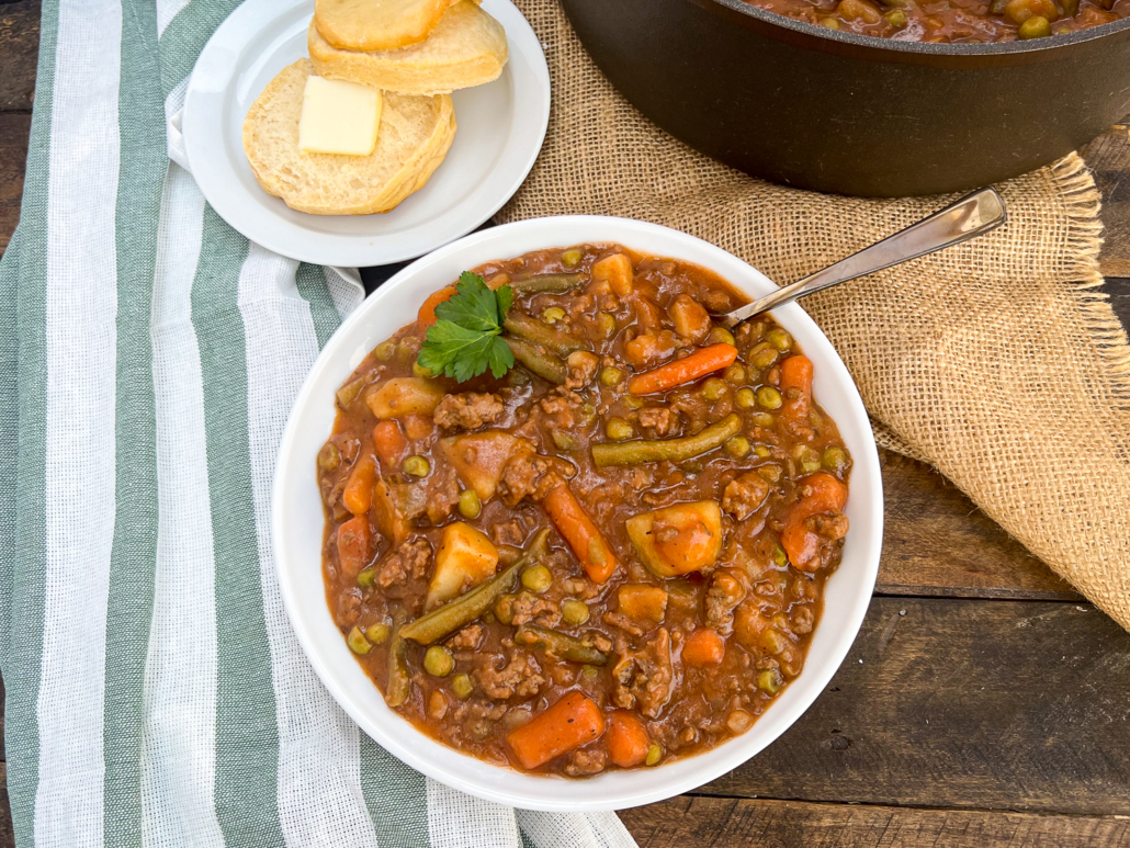 A bowl of ground beef stew in a white bowl served with a buttered biscuit.