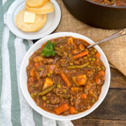 A bowl of ground beef stew in a white bowl served with a buttered biscuit.