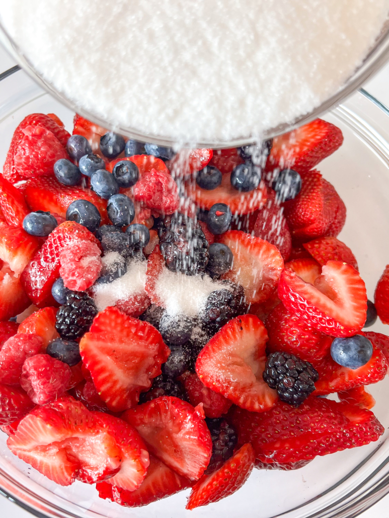 Mixed berries in a glass bowl with granulated sugar being poured on top.
